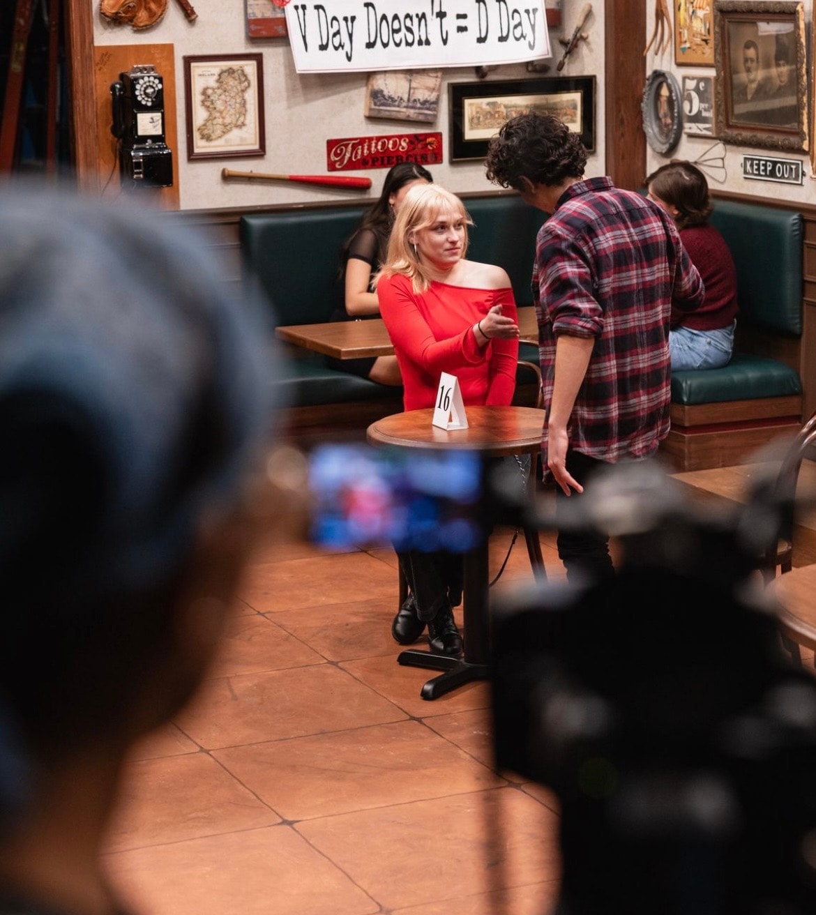 Lex Leone on camera in red off-shoulder top at diner table with co-star
