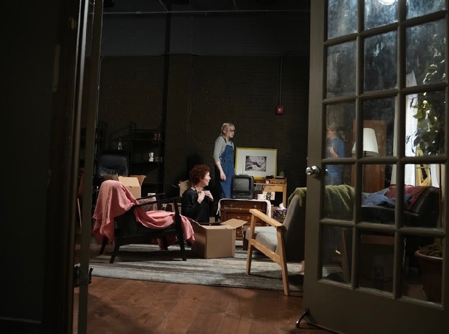 Lex Leone on film set in overalls standing in cluttered apartment set