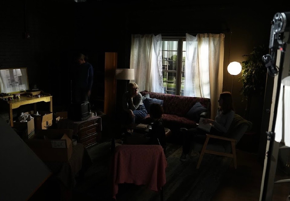 Lex Leone on film set seated on couch in dimly lit living room set