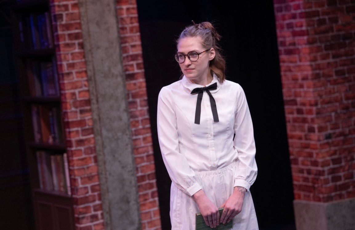 Lex Leone on stage in white blouse standing alone by brick wall
