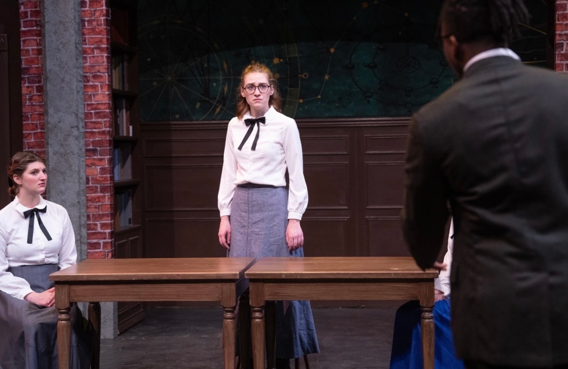 Lex Leone on stage in white blouse standing at desk facing co-star