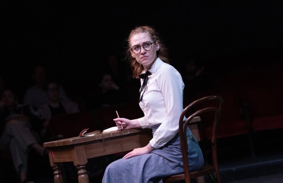 Lex Leone on stage seated at desk with pencil looking toward audience
